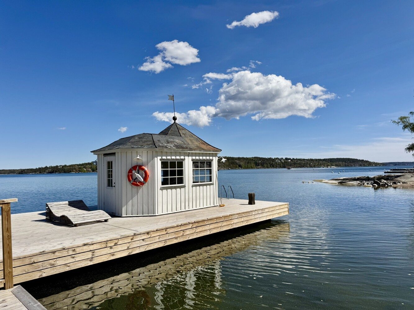 A traditional red-painted Swedish timber boathouse on a wooden jetty over calm, still water, with a small boat alongside.