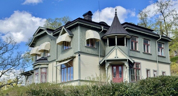 A large, ornate Swedish timber villa with decorative wooden detailing, set among tall trees.