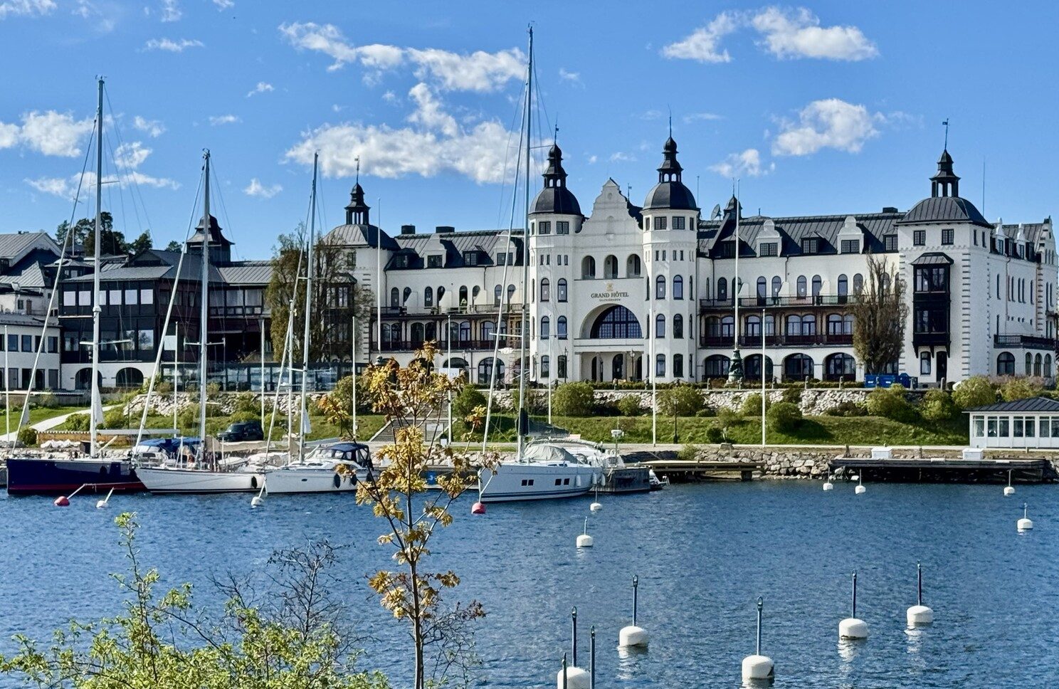 A grand white waterfront building with turrets and balconies behind a marina of moored boats under a clear blue sky in Sweden.